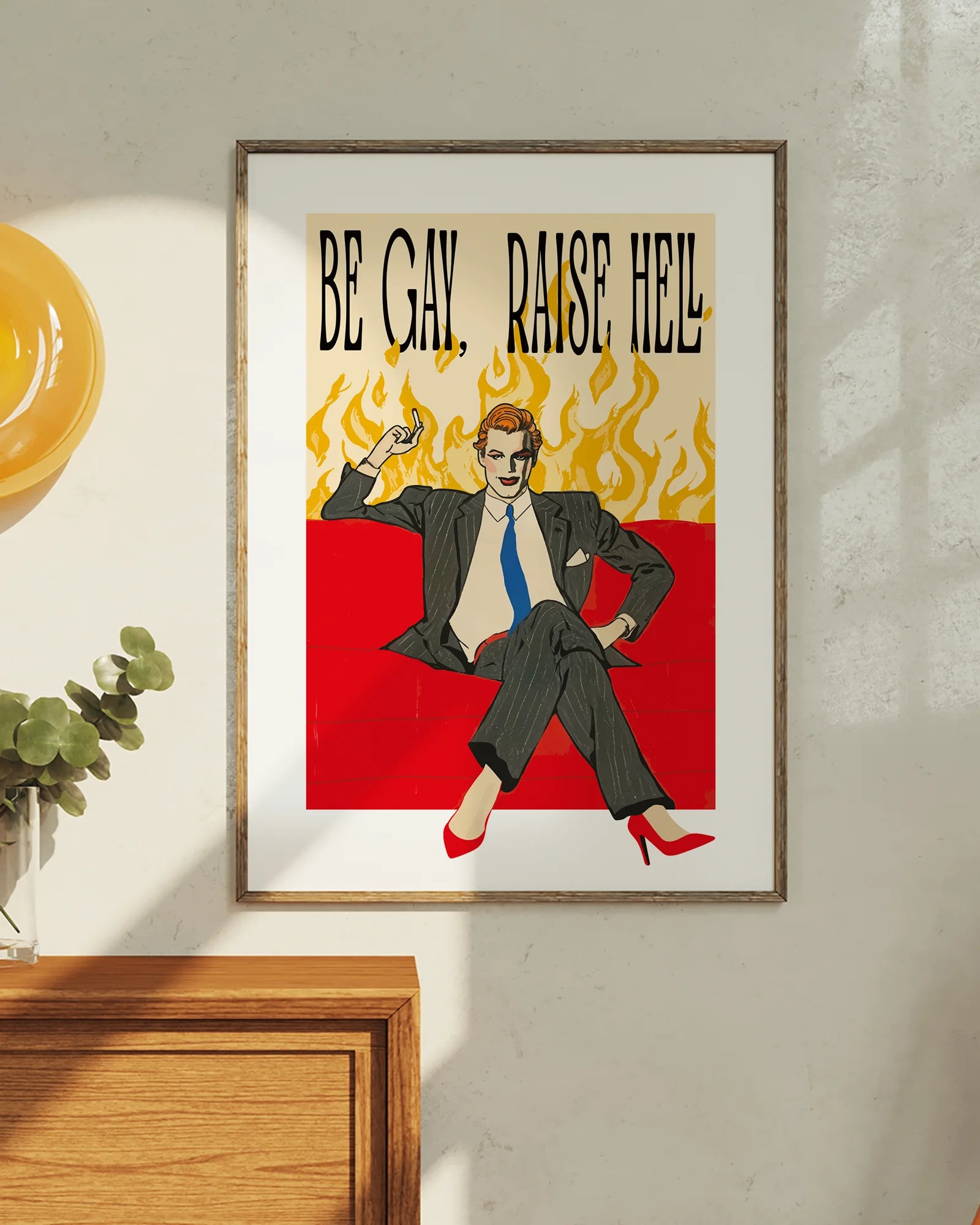 A framed poster featuring a man in make up, red high heels, sitting on a couch smoking, surrounded by massive flames, with the text "BE GAY, RAISE HELL".
