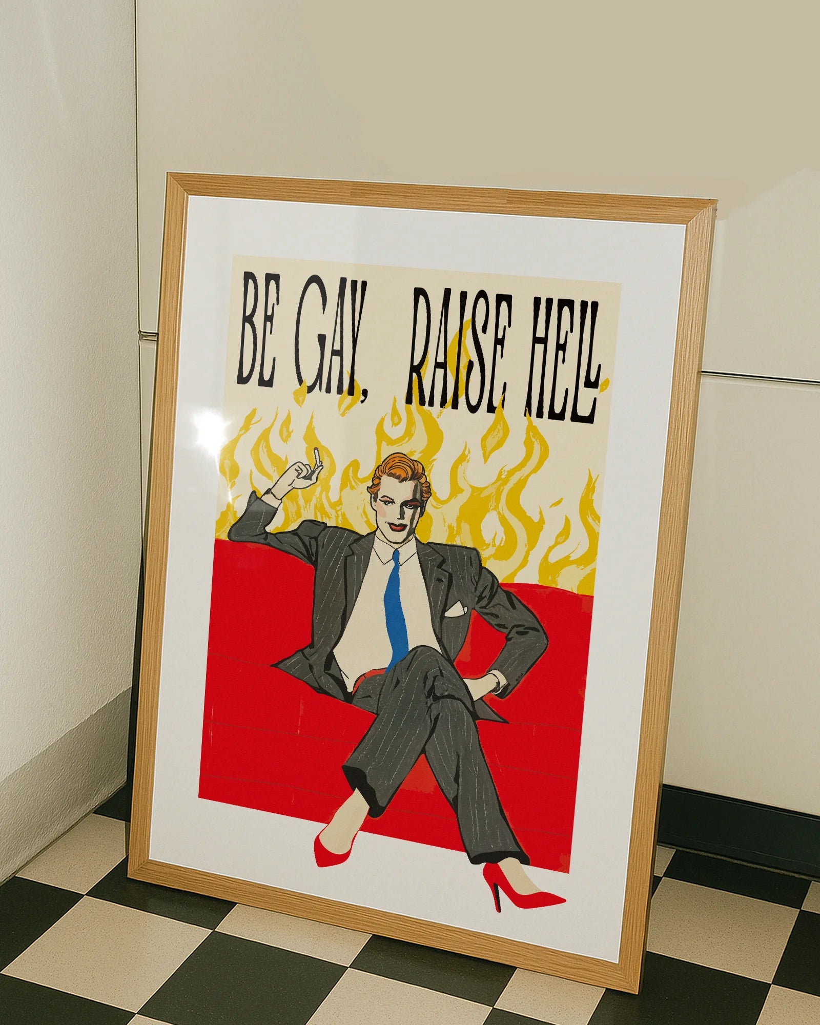 A framed poster featuring a man in make up, red high heels, sitting on a couch smoking, surrounded by massive flames, with the text "BE GAY, RAISE HELL".