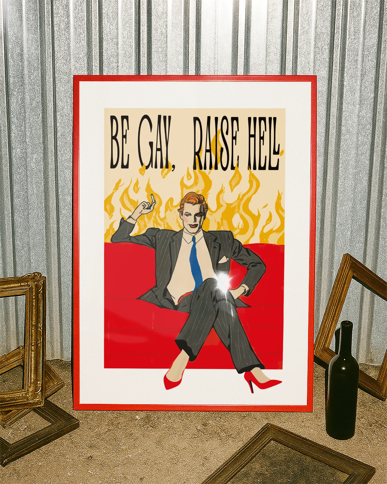 A framed poster featuring a man in make up, red high heels, sitting on a couch smoking, surrounded by massive flames, with the text "BE GAY, RAISE HELL".