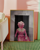 A poster with a muscular drag queen in a sparkling dress, full makeup, and wig, confidently strutting out of the ladies’ room.