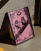 A poster featuring a female devil sitting on the edge of a coffin, while a female vampire sits inside. They hold each other’s hands romantically, creating a mysterious and intimate atmosphere.