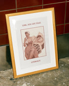 A poster featuring a woman wiping her mouth with a cloth napkin while sitting on the edge of a bed, with another woman lying behind her. Text on the artwork reads: “GIRL, YOU ATE THAT. LITERALLY.”