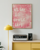 A framed poster with the text 'BE GAY, DO OTHER GAYS.', against a pink, abstract background.