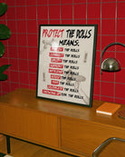 A framed print featuring the text “PROTECT THE DOLLS MEANS:” followed by:
employ the dolls
connect the dolls
uplift the dolls
support the dolls
befriend the dolls
house the dolls
feed the dolls
celebrate the dolls
showing up for the dolls