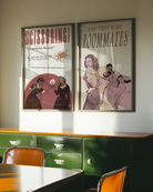 A framed poster depicting a woman lying in bed, and another woman undressing to join her — with the text “AND THEY WERE ROOMMATES.”