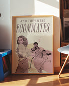 A framed poster depicting a woman lying in bed, and another woman undressing to join her — with the text “AND THEY WERE ROOMMATES.”