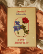 A framed poster featuring violets and red roses with the text:
"Roses are red, Violets are blue. I'm very gay, And so are you, boo."