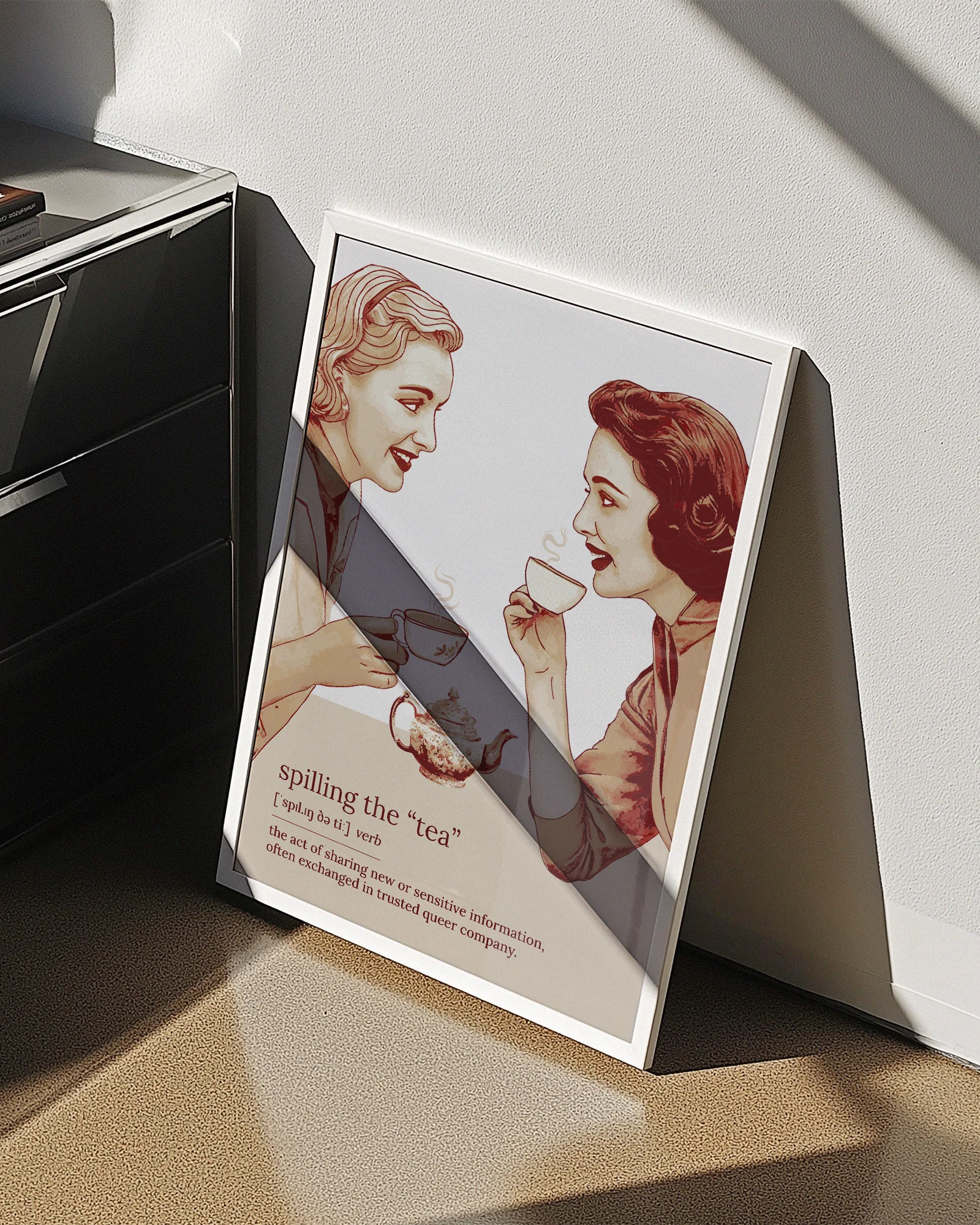 A framed poster featuring two women gossiping over tea, accompanied by the text: “Spilling The Tea – Verb – The act of sharing new or sensitive information, often exchanged in trusted queer company.”