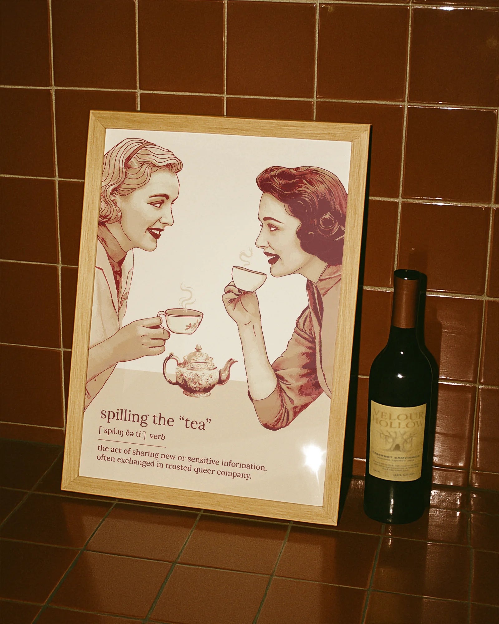 A framed poster featuring two women gossiping over tea, accompanied by the text: “Spilling The Tea – Verb – The act of sharing new or sensitive information, often exchanged in trusted queer company.”