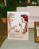 A framed poster featuring two women gossiping over tea, accompanied by the text: “Spilling The Tea – Verb – The act of sharing new or sensitive information, often exchanged in trusted queer company.”